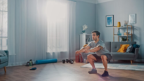 A man performs a squat exercise in the hardwood paneled, living room of his apartment. 