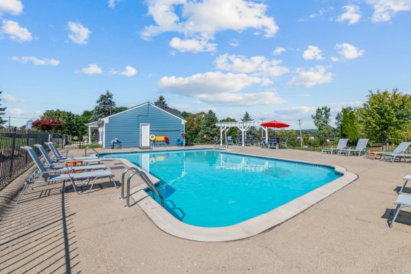Outdoor swimming pool with lounge chairs, blue sky, and green trees in the background on a sunny day.