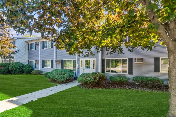 Two-story grey building with green lawn and trees, entrance door centered under sunny blue sky.