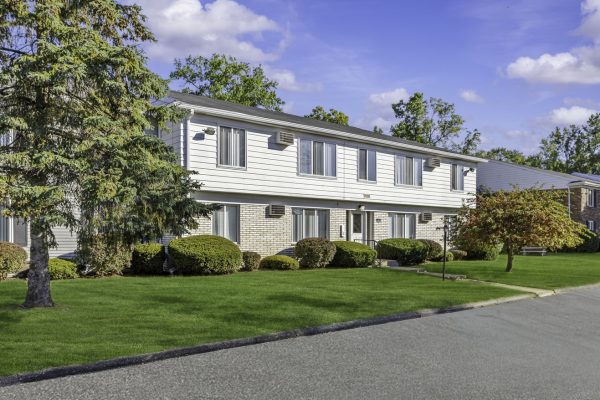 Two-story suburban apartment building with well-maintained lawn and trees under a clear blue sky.