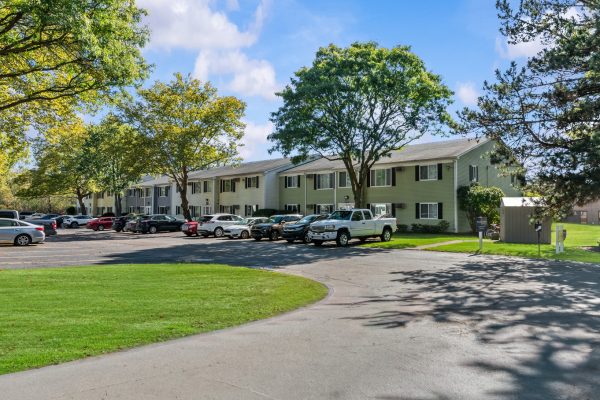 Apartment complex with parking lot, surrounded by trees under a clear blue sky.