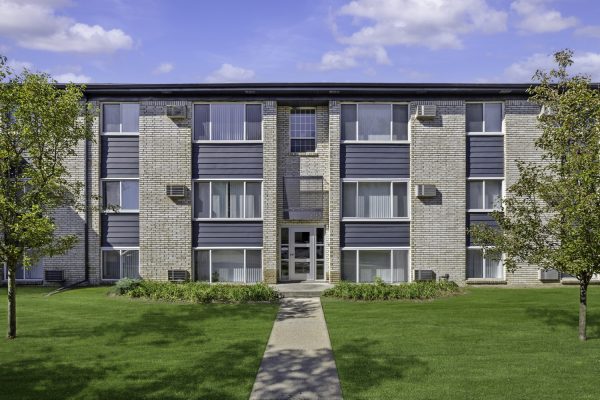 Three-story apartment building with modern facade, surrounded by green lawn and trees under a blue sky.