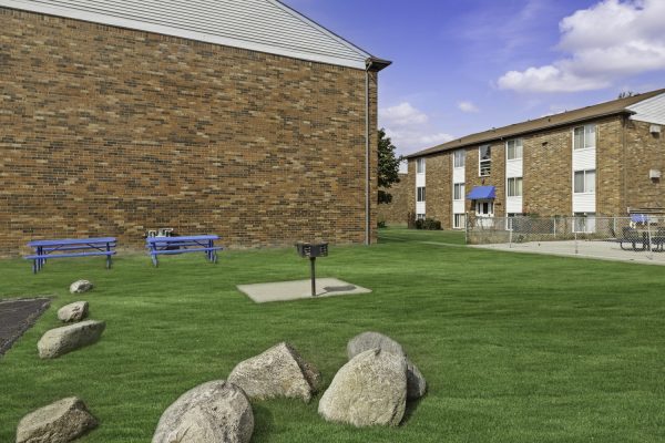 Apartment courtyard with picnic tables, grill, and green lawn under a blue sky.