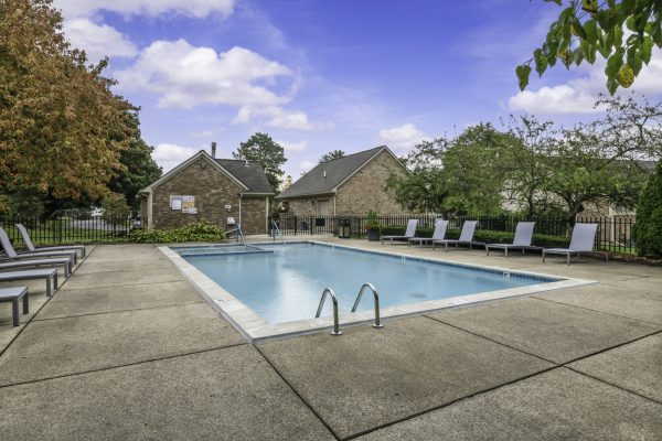 Outdoor swimming pool with lounge chairs, surrounded by trees, under a blue sky at a residential complex.