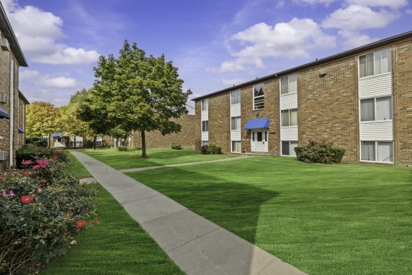 Apartment complex with brick buildings, green lawns, and a tree-lined pathway on a sunny day.