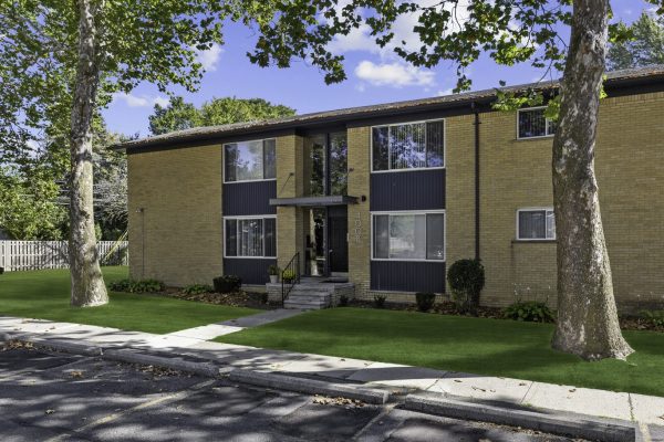 Two-story brick apartment building with trees and manicured lawn, bright sunny day.