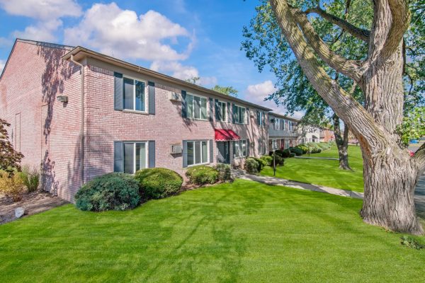 Brick apartment building with lush green lawn and large tree under blue sky.