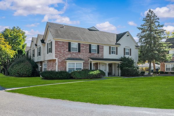 Large suburban house with brick facade, lush green lawn, and clear blue sky in a quiet neighborhood setting.