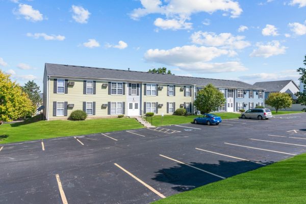 Apartment complex with grassy lawn and parking lot, featuring a blue sky backdrop and several parked cars.