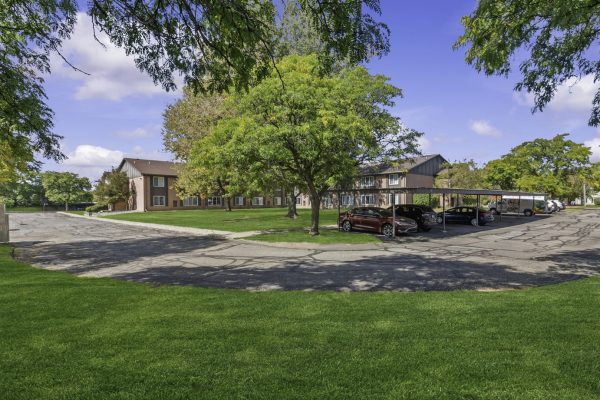 Apartment complex with parking lot, green grass, and tree-lined road under a clear blue sky.