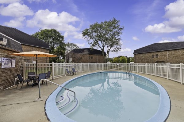 Outdoor oval pool with fence, patio chairs, and an orange umbrella on a sunny day.