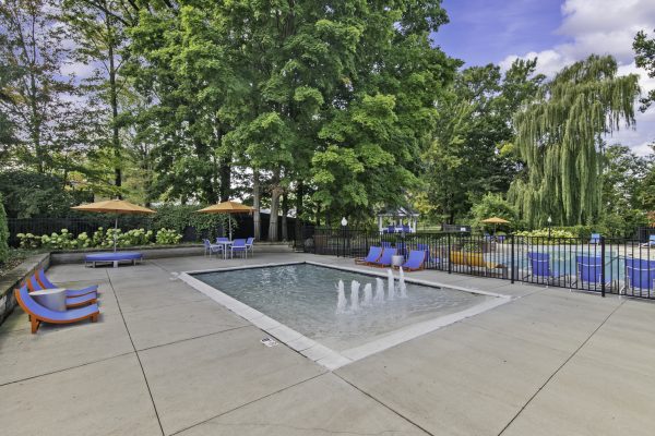 Outdoor swimming pool with fountains, sun loungers, and umbrellas surrounded by lush greenery and trees under a blue sky.