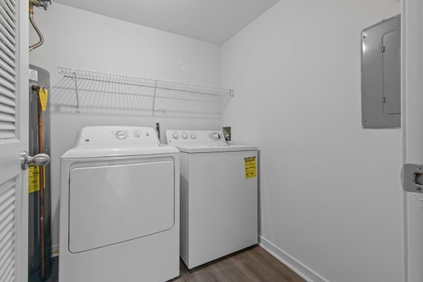 White laundry room with washer, dryer, water heater, and wire shelf in a small, functional space.