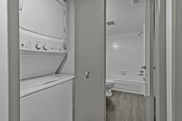 Compact laundry and bathroom area with stackable washer-dryer, white bathtub, and wooden floor.