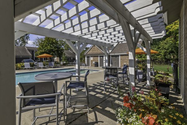 Poolside patio with chairs under a pergola, surrounded by colorful flowers and umbrellas in a sunny outdoor setting.