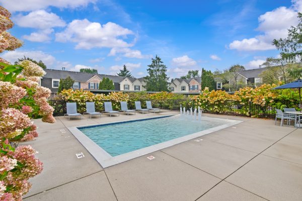 Apartment complex with a landscaped outdoor pool, lounge chairs, and flowers under a blue sky.