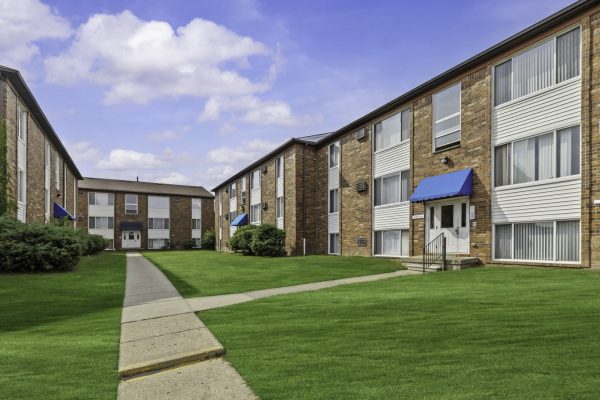 Brick apartment buildings with blue awnings, surrounded by lush green lawns under a clear blue sky.