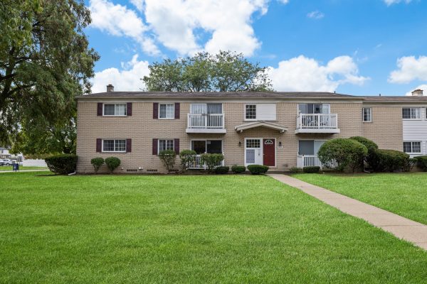 Two-story beige brick apartment building with balconies, surrounded by green lawn under a partly cloudy sky.