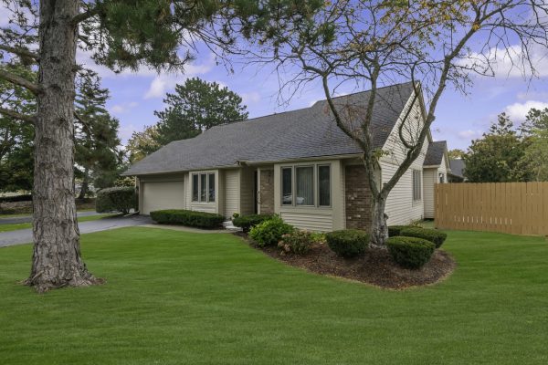 Single-story suburban home with manicured lawn, surrounded by trees and a wooden fence under a blue sky.