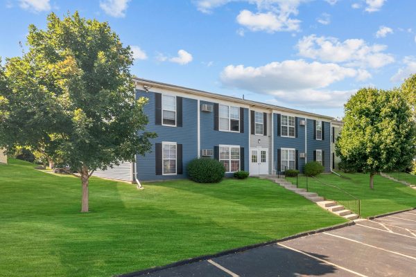 Blue two-story apartment building with lush green lawn and trees under a clear sky.