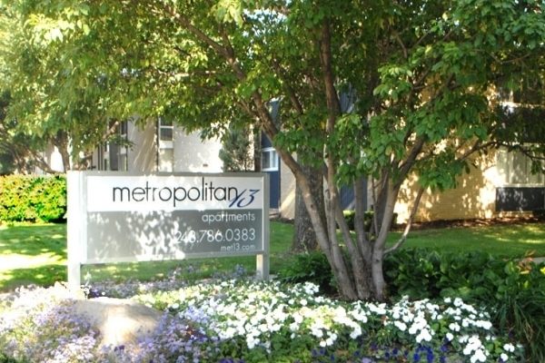 Sign for Metropolitan 13 Apartments surrounded by lush greenery and flowers in a sunny garden setting.