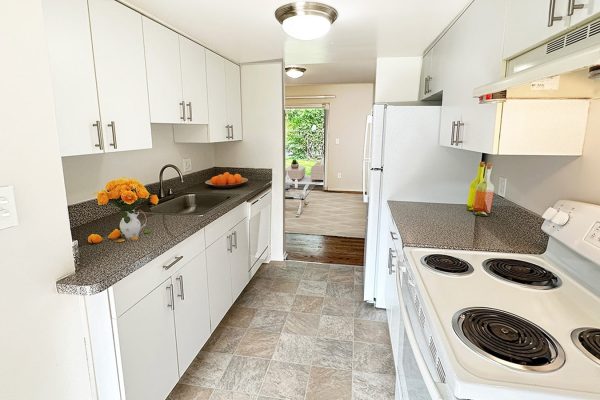 Modern kitchen with white cabinets, granite countertops, and a view into a bright living area through a doorway.