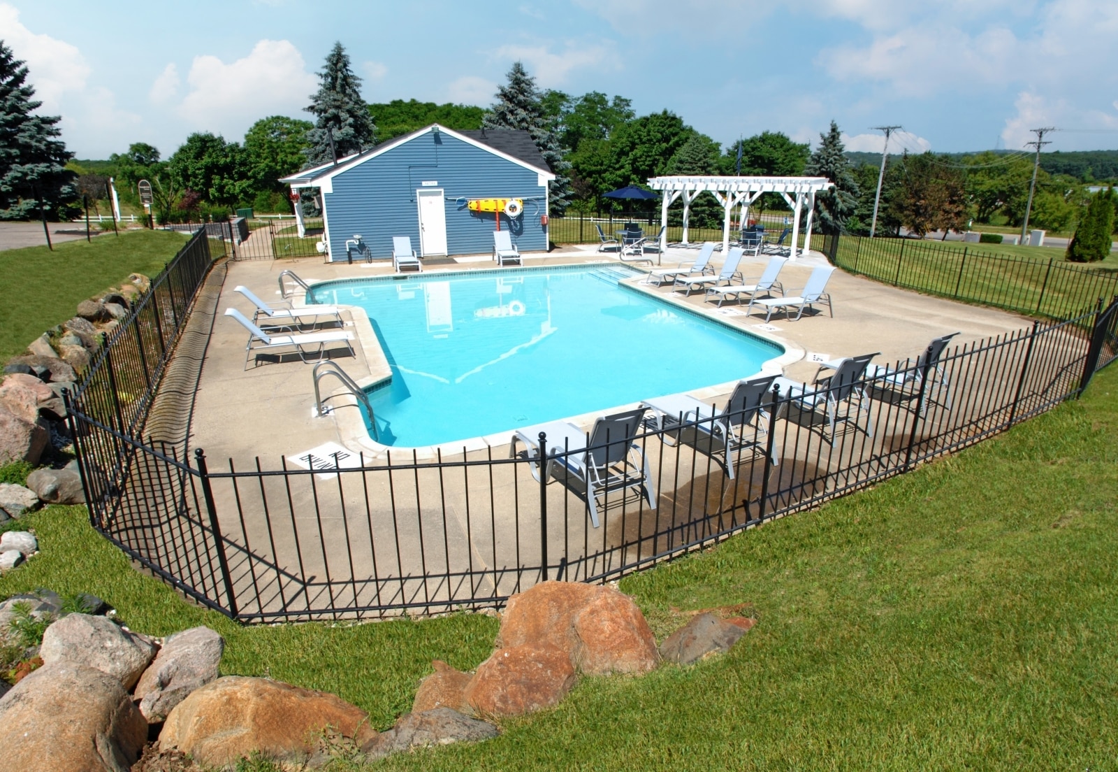 Fenced outdoor swimming pool with lounge chairs and a blue building in a sunny, grassy setting.