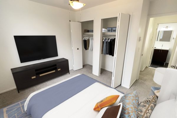 Modern bedroom with TV, open closet, and bathroom; stylish decor with blue bedspread and decorative pillows.
