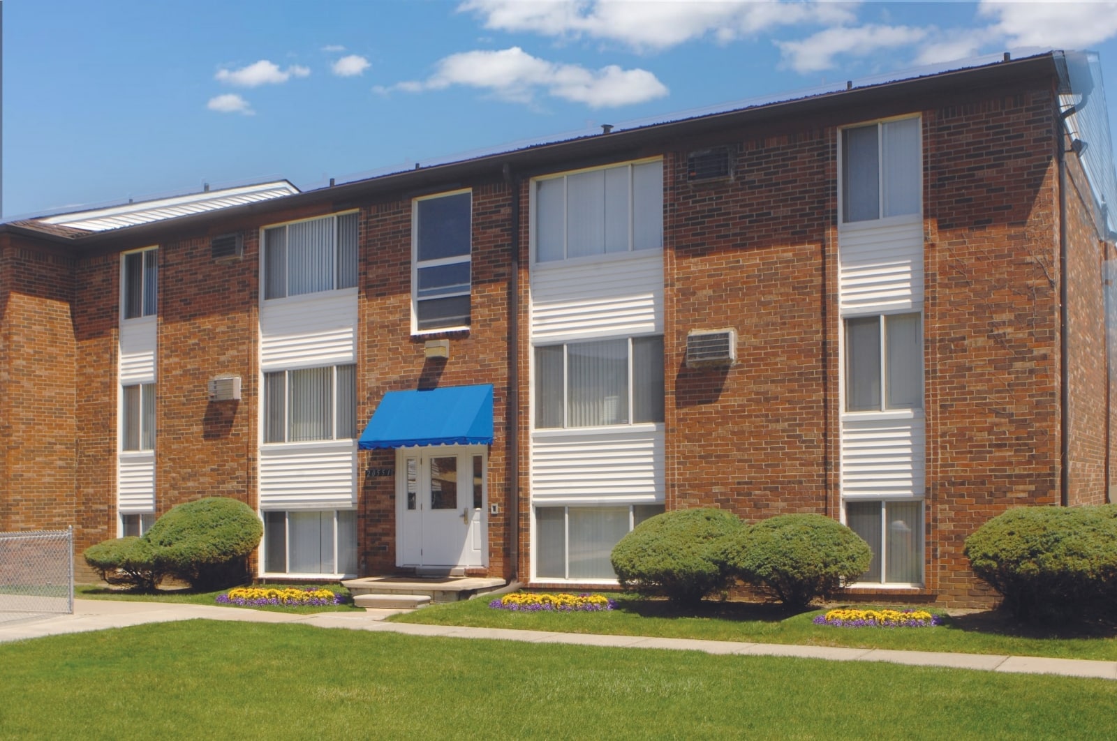 Three-story brick apartment building with blue awning, manicured bushes, and clear skies.